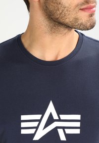 Tamsiai mėlynas medvilninis marškinėlis su apvalia iškirpte, ant priekinės pusės turintis didelį baltą logotipą su kampuotu dizainu ir dryželiais.