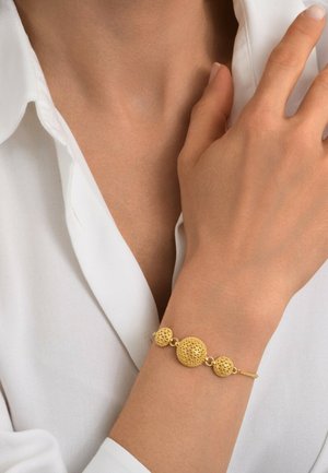 Bracelet en or avec trois éléments sphériques texturés, reliés par une chaîne délicate, porté sur un poignet nu contre une blouse blanche.