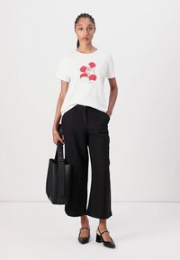Witte t-shirt met rode bloemenillustratie, gecombineerd met zwarte wijde broek. Model houdt een zwarte leren tote bag vast en draagt zwarte hakken.