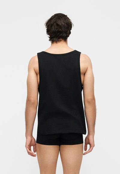 Άνδρας που φοράει μαύρο αμάνικο τοπ και μαύρο σορτς, στέκεται με την πλάτη του σε λευκό απλό φόντο.