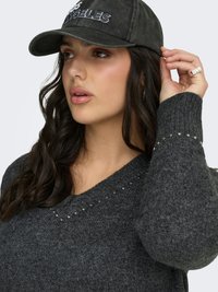 Kvinna med långt mörkt hår som bär en mörkgrå nitsweater och en svart keps med texten "Los Angeles", justerar kepsen med handen.