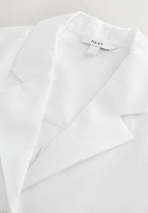Biely blejzer s výstrihom s výrezom, hladkou textúrou a viditeľnou nálepkou s nápisom "NEXT." Má strihané klopy a čistý dizajn.