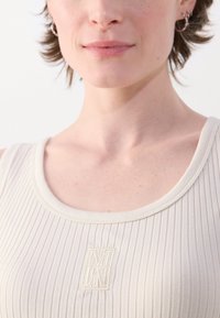 Canottiera rigata beige chiaro con scollatura rotonda. Presenta un tessuto testurizzato e un piccolo logo ricamato al centro.