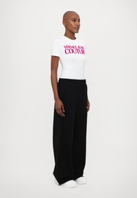 Balta īsām piedurknēm blūze ar uzrakstu "Versace Jeans Couture" rozā krāsā, apvienota ar brīvām melnām sporta biksēm un baltām kedām.