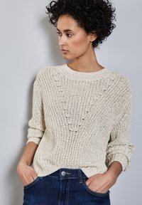 Maglione beige lavorato a maglia con una vestibilità ampia, caratterizzato da un colletto tondo e polsini arrotolati. Indossato con jeans blu scuro, che mostrano una trama evidente.