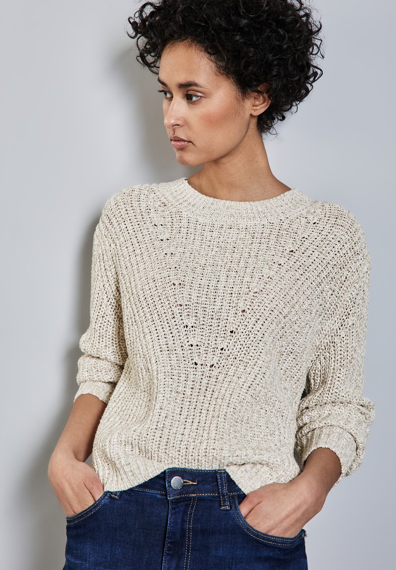 Maglione beige lavorato a maglia con una vestibilità ampia, caratterizzato da un colletto tondo e polsini arrotolati. Indossato con jeans blu scuro, che mostrano una trama evidente.