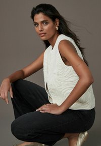 Gilet matelassé blanc avec des motifs en relief, décolleté en V profond et sans manches, associé à un jean noir et des chaussures métalliques dorées.