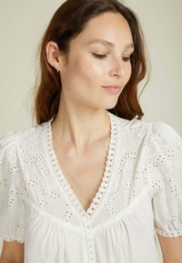 Blouse en coton blanc avec des motifs floraux brodés, décolleté festonné et manches courtes bouffantes. Texture douce avec une coupe décontractée.