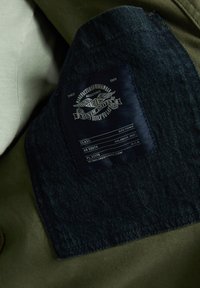 Denim label with blue fabric and white text against an olive green background; features branding and product details; stitched edges.