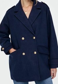 Donna che indossa un cappotto doppiopetto blu navy con bottoni dorati, una mano in tasca, abbinato a jeans blu e una collana con pendente dorato.
