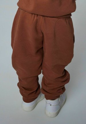 Bruine katoenen sweatpants met een elastische tailleband en boorden, gecombineerd met witte sneakers tegen een lichte achtergrond. Relaxte pasvorm en eenvoudig ontwerp.