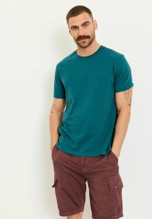 Mann mit Schnurrbart und Tattoos, der ein türkisfarbenes T-Shirt mit kurzen Ärmeln und kastanienbraune Cargo-Shorts trägt und mit den Händen in den Taschen vor einer weißen Wand steht.