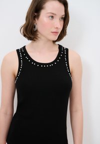 Zwarte mouwloze top met een ribbelstructuur, voorzien van zilveren kraaltjes langs de halslijn. Eenvoudig, aansluitend ontwerp zonder verdere versieringen.