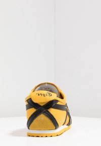 Basket jaune avec des rayures noires croisées vue de dos sur une surface blanche contre un mur blanc.