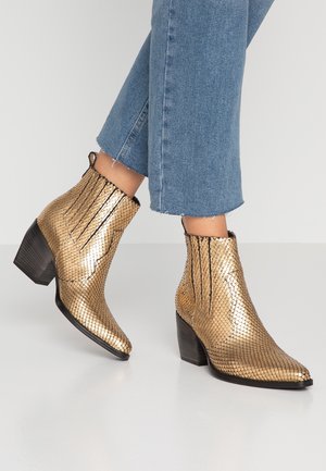 Kotníkové boty s kovovým zlatým texturováním, špičatými špičkami a blokovými podpatky, nošené s odhalenými modrými džíny na jednobarevném pozadí.