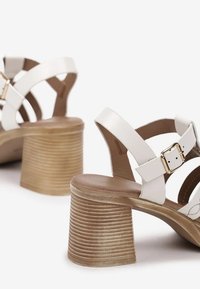 Sandales à talons blancs avec des brides de cheville ajustables, une boucle circulaire dorée et des talons en bois rainuré. Doublure intérieure lisse et design minimaliste.