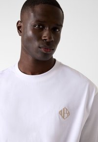 Biała bawełniana koszulka z okrągłym dekoltem, z małym haftowanym beżowym logo na lewej stronie klatki piersiowej. Gładka faktura, luźny krój.