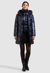 Abrigo de plumón azul marino con un cuello alto, textura acolchada, silueta ajustada y cremallera frontal. Llevado sobre una falda negra de canalé y botas negras hasta la rodilla.