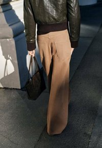 Bruine wijde broek met een gladde textuur, gecombineerd met een nauwsluitende donkerleren jas en een bruine tote bag, gezien van achteren.