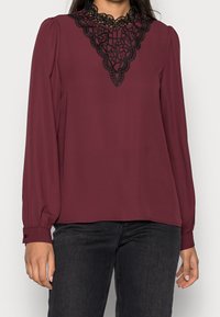 Mörkröd blus med långa ärmar, med en svart spetsdetalj vid halslinningen. Slätt tyg med avslappnad passform och knäppta manschetter.