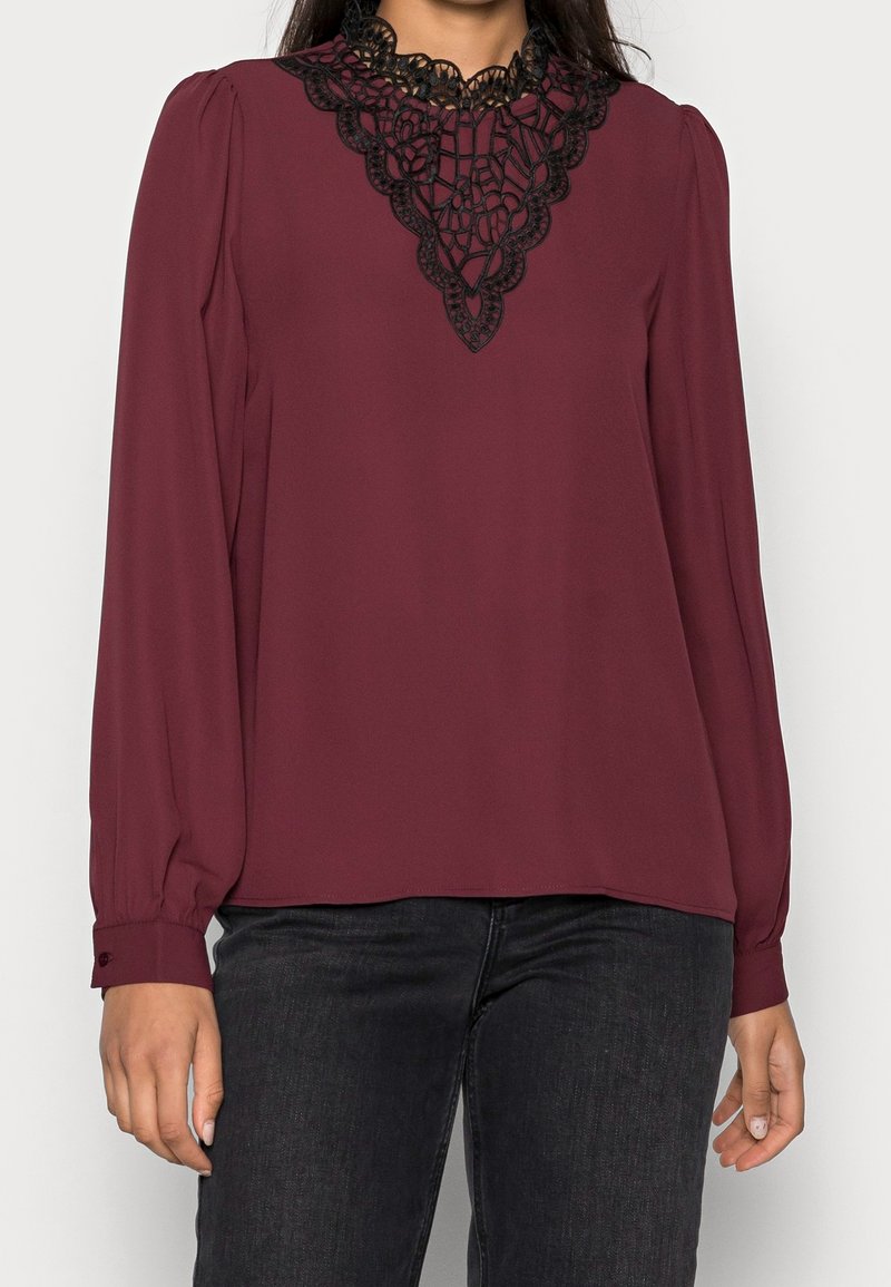 Mörkröd blus med långa ärmar, med en svart spetsdetalj vid halslinningen. Slätt tyg med avslappnad passform och knäppta manschetter.