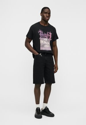 Μαύρο γραφικό t-shirt με φλογέρες πάνω σε κεντρικό σχέδιο, συνδυασμένο με μαύρο τζιν σορτς και μαύρα αθλητικά παπούτσια. Ορατές λευκές κάλτσες αστραγάλου.