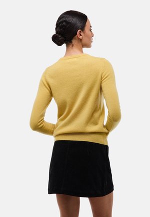 Femme aux cheveux noirs en chignon portant un pull jaune moutarde et une jupe noire, vue de dos sur un fond clair uni.