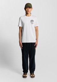 Weißes T-Shirt mit kurzen Ärmeln und einem schwarzen Löwen-Motiv, kombiniert mit navyfarbenen Hosen und braunen gemusterten Schuhen, akzentuiert durch eine olivfarbene Mütze.