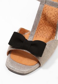 Sandale ouverte argentée avec une texture pailletée, un nœud en tissu noir sur la sangle avant et une semelle intérieure beige à motifs.