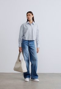 Φωτεινό μπλε πουκάμισο με ρίγες και κουμπιά, ψηλόμεσα ξεθωριασμένα τζιν, λευκά αθλητικά παπούτσια και μια κρεμ τσάντα ώμου με ομαλή υφή.