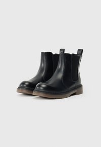 Des bottes Chelsea en cuir noir avec des panneaux élastiques sur les côtés, un bout rond et une semelle en caoutchouc texturée avec un bord marron. Tirants à l'arrière.