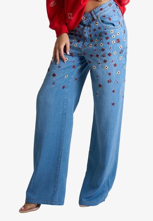 Jeans blu a gamba larga con stelle e quadrati ricamati rossi e gialli sparsi, indossati con un top rosso e sandali.
