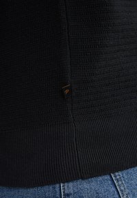 Black knitted sweater featuring a textured pattern with ribbed hems. Small branded label stitched at the side, paired with blue denim jeans.