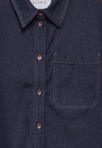 Camicia in denim scuro con colletto a punta, due bottoni marroni e taschino sul petto con dettagli di cucitura visibili.