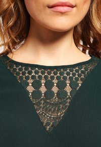 Haut vert foncé avec un col en V orné de détails en dentelle complexes, incluant des motifs circulaires et une texture de tissu lisse.