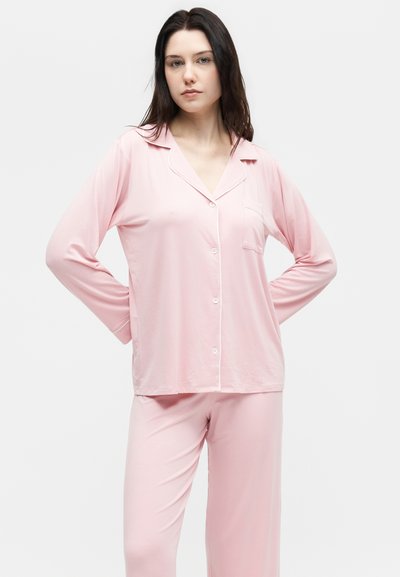 Γυναίκα στέκεται με τα χέρια πίσω από την πλάτη φορώντας ανοιχτό ροζ πουκάμισο με μακριά μανίκια και κουμπιά και παντελόνι σε ίδια απόχρωση, σε απλό λευκό φόντο.