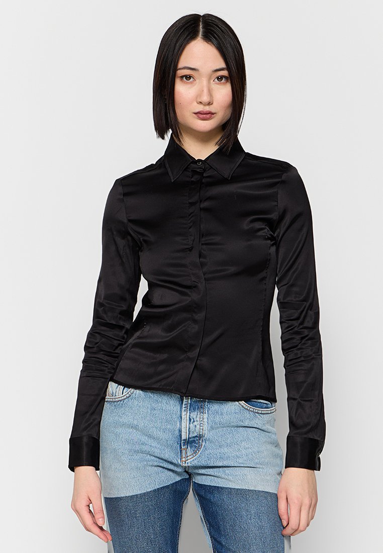 Schwarzes Langarmshirt aus glattem Satin; mit einem kragenartigen Ausschnitt, vorderem Knopfverschluss und taillierter Passform. Kombiniert mit Jeans.