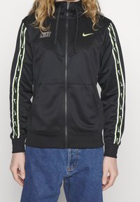 Černá bunda na zip z hladké tkaniny, s zelenými a bílými pruhy na rukávech, s kapucí a logem na hrudi.