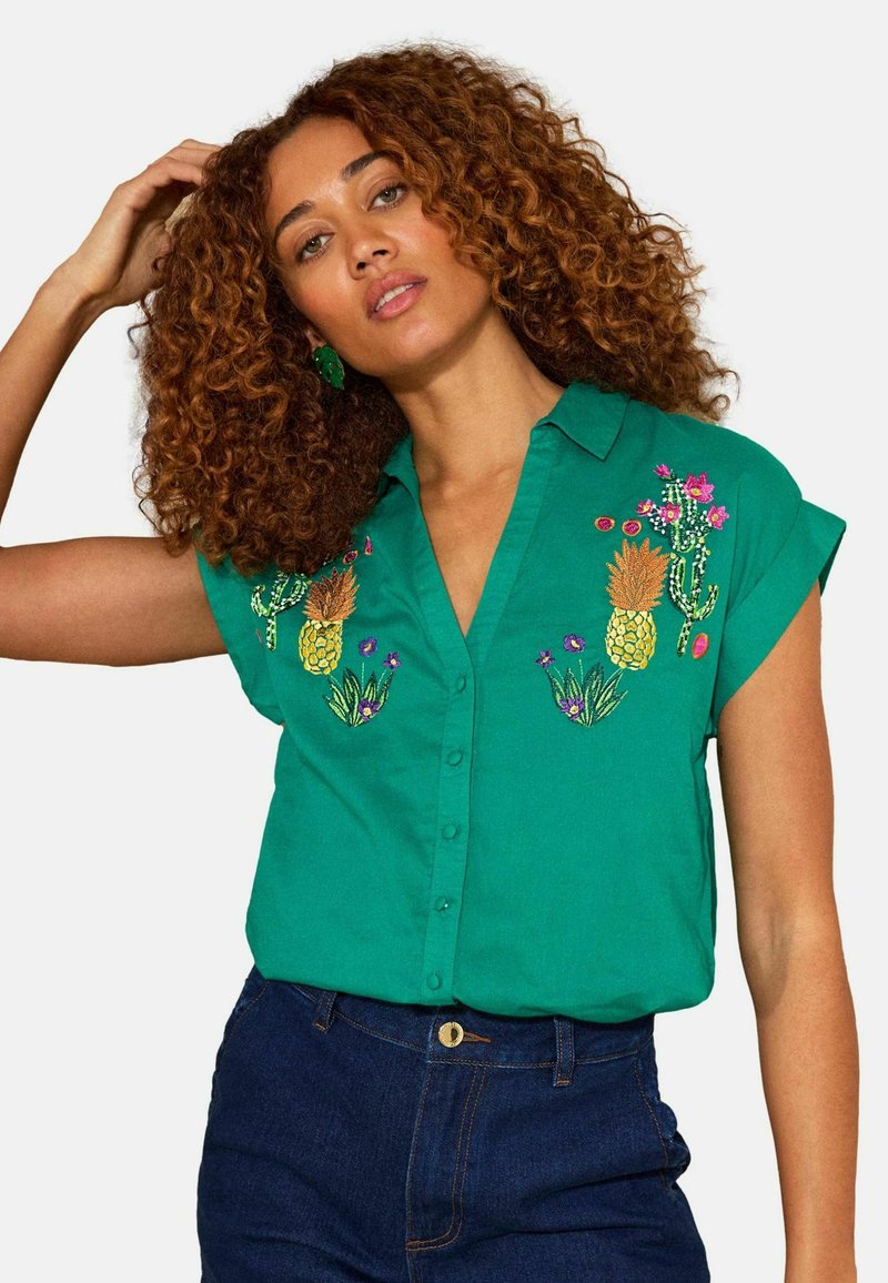 Femme aux cheveux bouclés portant une chemise verte à manches courtes avec des boutons et des broderies d'ananas et de cactus, associée à un jean bleu foncé.