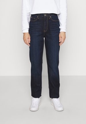 Person trägt dunkelblaue Jeans mit geradem Bein, weißes Langarmshirt und weiße Turnschuhe, steht vor einem schlichten hellen Hintergrund.