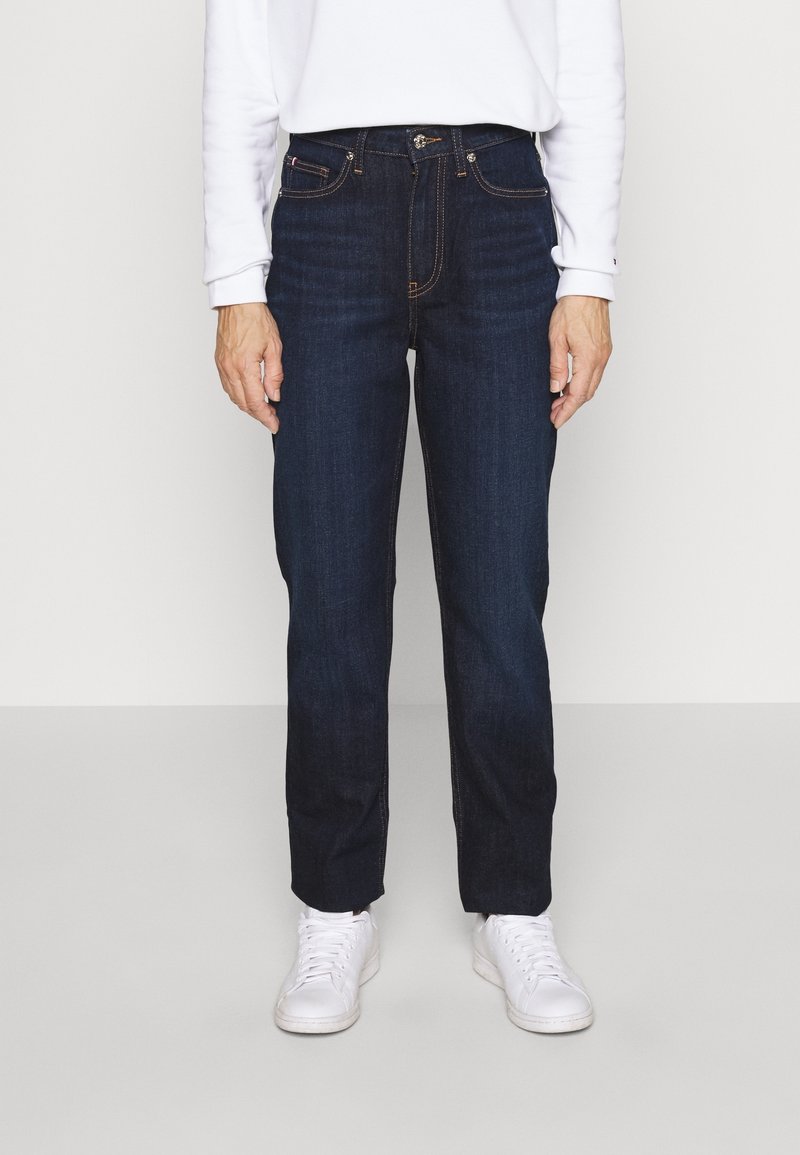 Person trägt dunkelblaue Jeans mit geradem Bein, weißes Langarmshirt und weiße Turnschuhe, steht vor einem schlichten hellen Hintergrund.