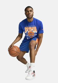 Kék grafikus póló narancssárga részletekkel és tigris mintával, kék csíkos rövidnadrággal, fehér zoknikkal és fehér/piros kosárlabdicipővel párosítva.