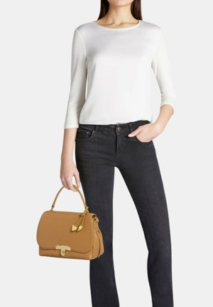 Donna con una maglia bianca a maniche lunghe e jeans neri che tiene una borsa in pelle color cuoio con ciondoli a forma di cuore.