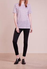 Bílý tričko s fialovými diagonálními pruhy a malou modrou grafikou na hrudi; v kombinaci s černými slim-fit kalhotami a lesklými černými botami.