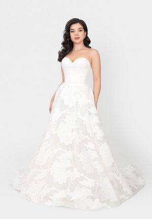 Donna con capelli lunghi e scuri che indossa un abito da sposa bianco senza spalline con gonna in pizzo floreale, in piedi davanti a uno sfondo chiaro e semplice.