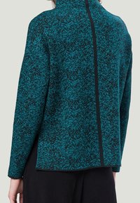 Haut texturé en teal et noir avec un col haut et une couture au dos. Le tissu présente un motif émaillé, créant une profondeur visuelle.