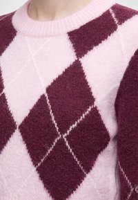 Gros plan d’un pull avec un motif losanges rose et rouge foncé et un col rond côtelé rose.