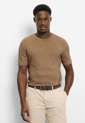 Man met kort krullend haar, gekleed in een bruin shirt met korte mouwen, beige broek en een donkere riem, staand tegen een witte achtergrond.