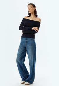 Ein marineblauer, schulterfreier, gerippter Pullover, kombiniert mit weiten blauen Jeans und beigen Sneakern, zeigt einen lässigen Schnitt und eine glatte Textur.