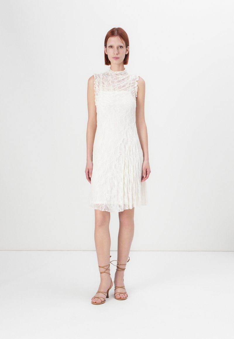 Vestido branco de renda com um colarinho alto, mangas em camadas e uma silhueta fluída, combinado com sandálias de tiras bege. Tecido liso e texturizado.